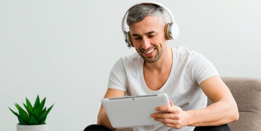 Aluno da graduação EAD sentado em um sofá com headphones, sorrindo enquanto olha para um tablet.
