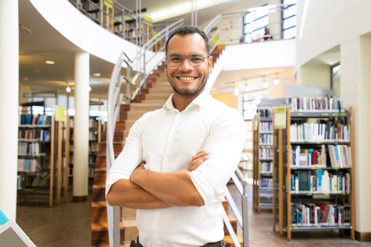 perfil-do-estudante-de-teologia-Homem negro de camisa branca está em pé, de braços cruzados, dentro de uma biblioteca.